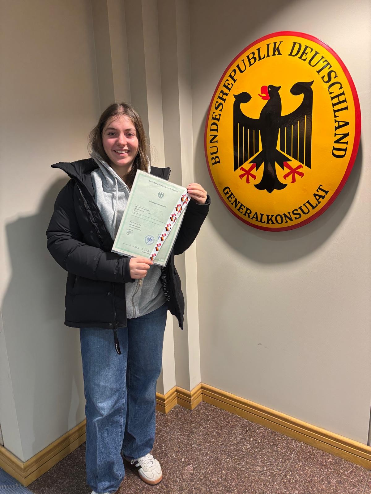 A woman stood in front of a german emblem on a wall whilst holding a certificate