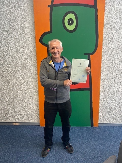 A man stood in front of a picture on a wall whilst holding a certificate